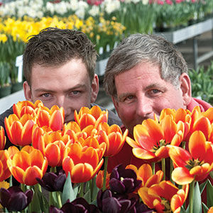 Two men hide the bottom halves of their faces behind a bunch of red and yellow tulips