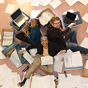 Three people sit among scattered papers on the ground and look up at the camera