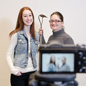Two women holding a piece of farm equipment stand in front of a camera sitting on a tripod so you can see the image on the camera at the same time