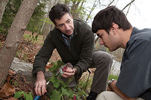 Two men crouch on the ground while one holds something in his hand out to the other