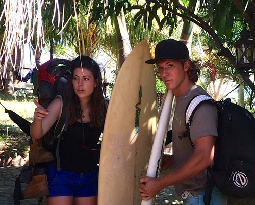 A man and a woman carry a surf board and backpacks outdoors