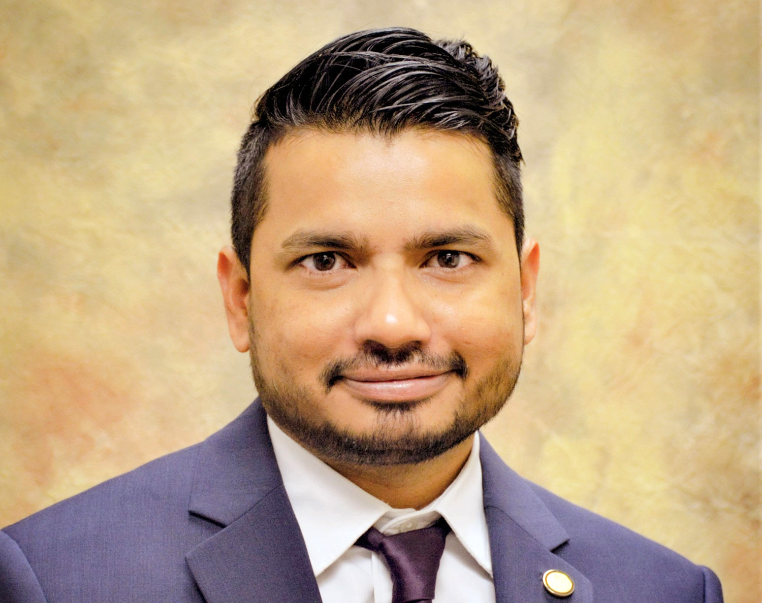 A headshot photo of Kiran Gadhave, wearing a blue suit, pastel shirt and navy blue tie, and smiling at the camera.