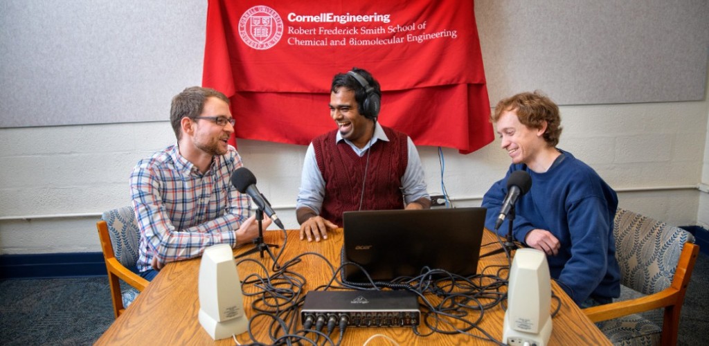 Students sit with podcast equipment in front of a Cornell Engineering flag