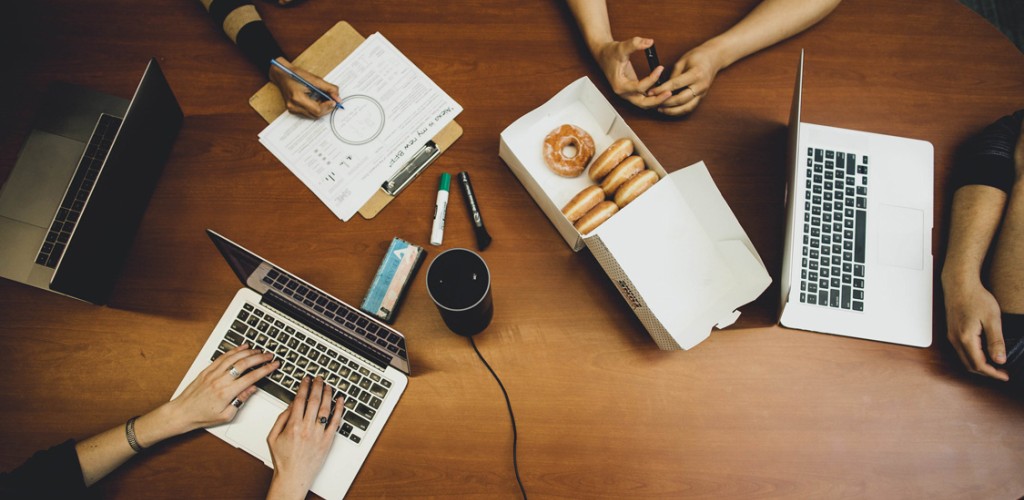 An arial view of men and women sitting at a table with laptops, paper and donuts, collaborating