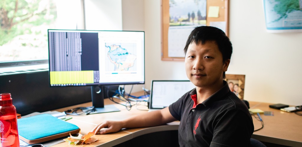 An asian male sitting at a desk in his office with a computer behind him