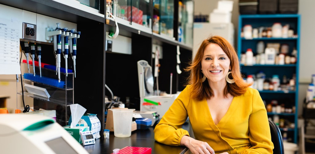 A Caucasian female with red hair in a yellow shirt sitting at a lab bench 