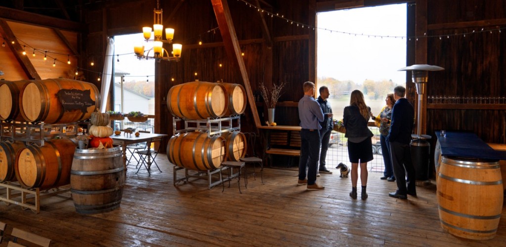 A group of adults standing and talking in a wooden barn containing wine barrel decor
