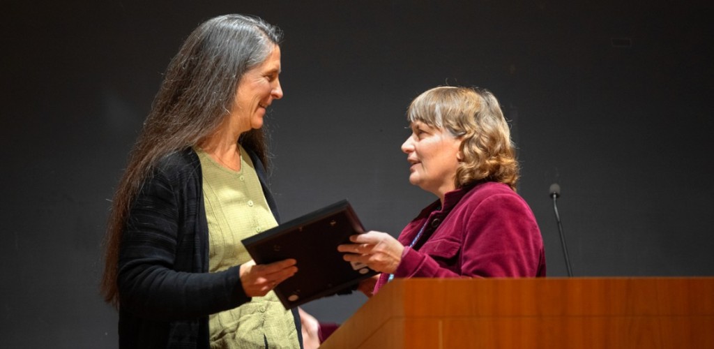 Two women facing each other standing on a stage in front of a podium with an award being passed off between them.  