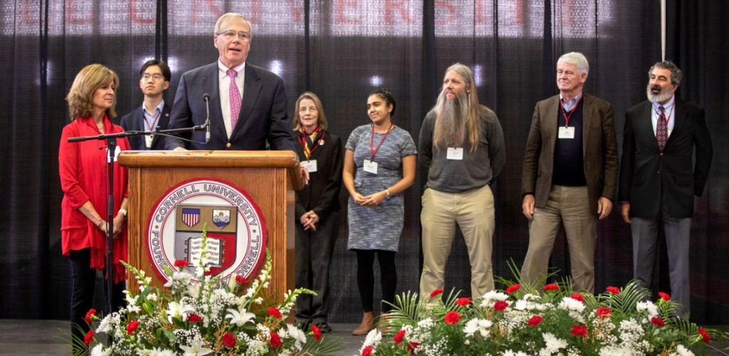 Men and women standing on a stage behind a man standing and speaking at a podium