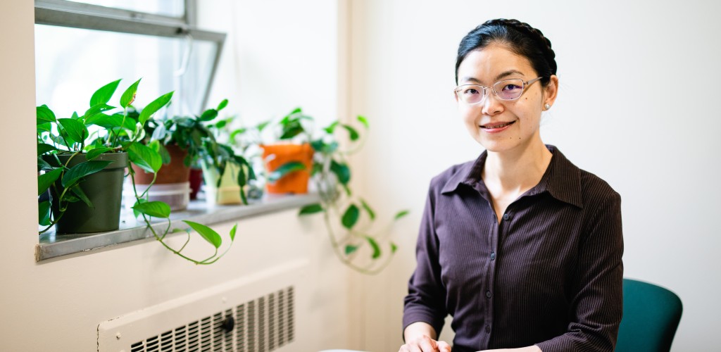 An asian female sitting at a table with her hands crossed. She is wearing glasses and a purple button down shirt and has her hair tied up in a braid. She is sitting in a white-walled office.
