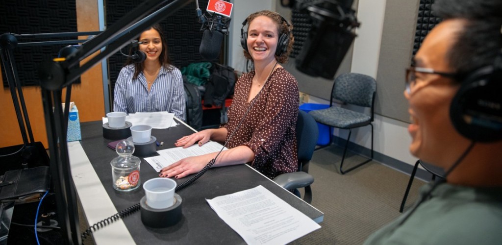 Two women and one male sitting at a table in front of microphones talking and laughing with one another