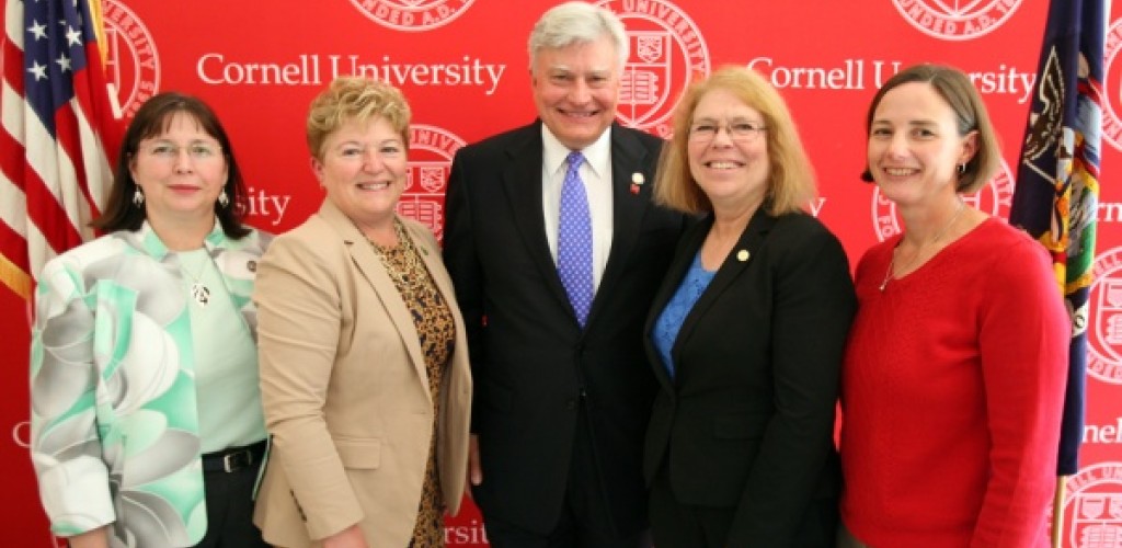 Four women and a man pose together for a photo in front of the Cornell seal