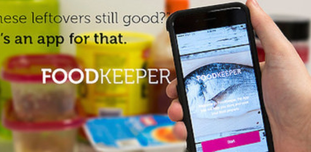 A hand holds an iPhone with the Foodkeeper application open on it in front of an ope fridge with containers of food in it
