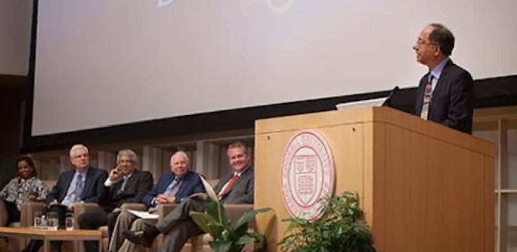 Five people sit on chairs on stage while one man stands at a podium and speaks