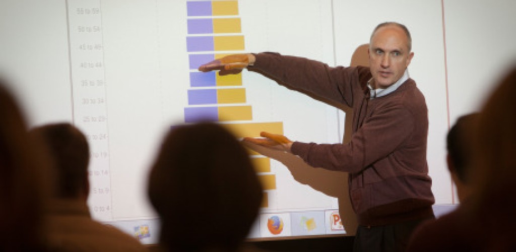 A man lectures in front of a class while pointing to the projection screen