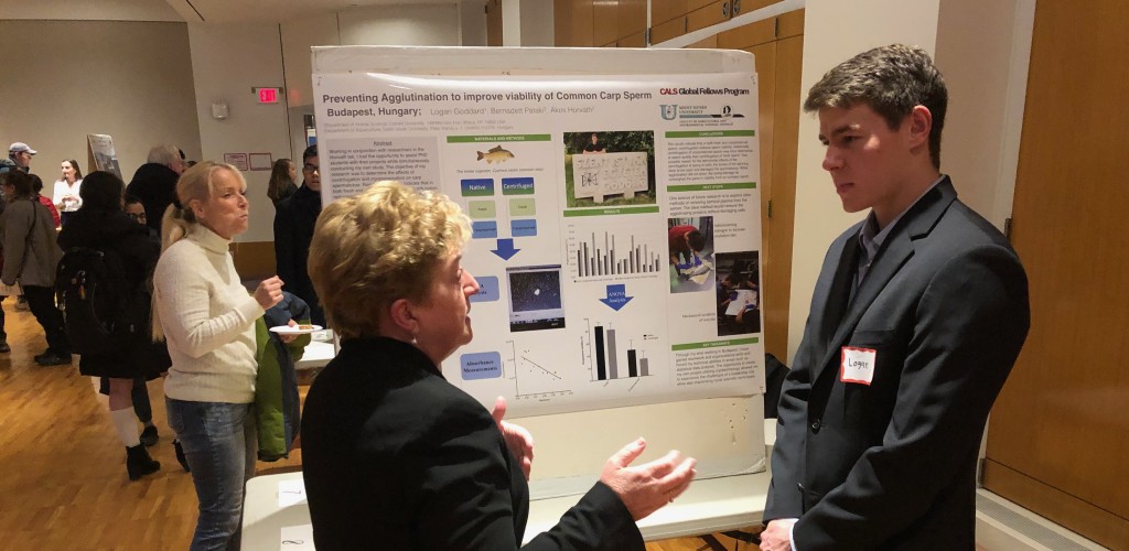 A woman talks to a young man standing in front of a poster that he made