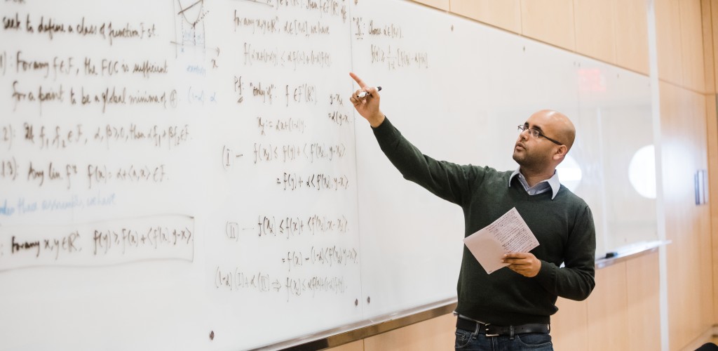 a bald man with glasses writes a complex equation on a whiteboard