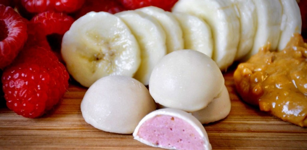 raspberries, a spoonful of peanut butter, sliced bananas and white smoothie bites on a wooden surface