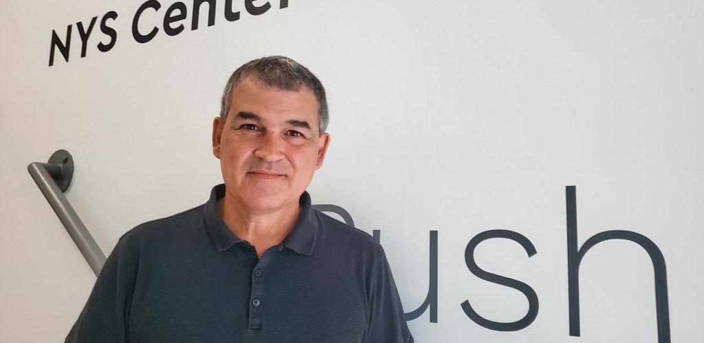 Headshot of Mark Scoville. Man standing on stairs with white background, wearing short-sleeved black polo shirt. 