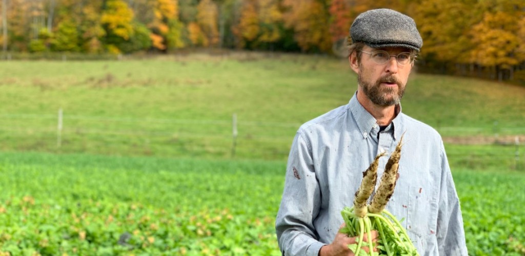 A man stands in a green field with two root vegetables in his hand.