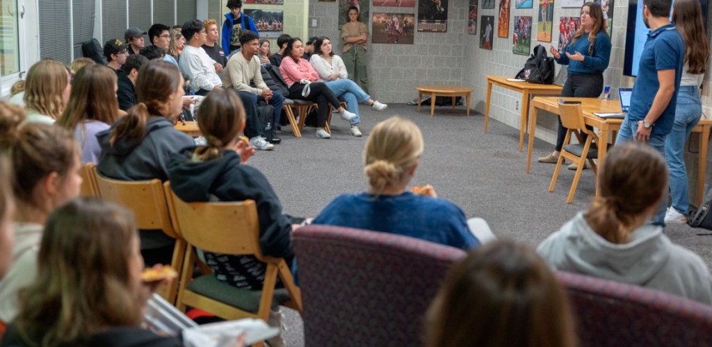 Students are seated in a half circle listening to a speaker.