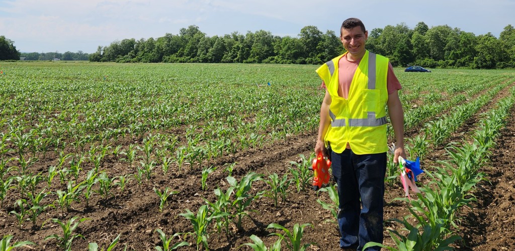 Sadiel standing in a farm field wearing a fluorescent vest and carrying research equipment.