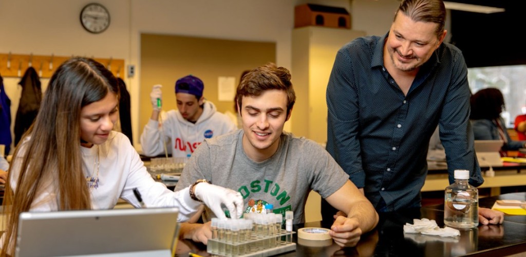 A female and male student work with a male teacher in a laboratory classroom holding vials. 