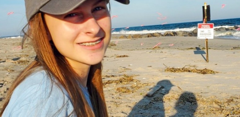A girl in a hat holding binoculars sits on a beach.