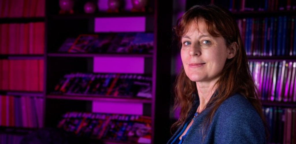 A woman stands in front of a bookshelf washed in purple light.