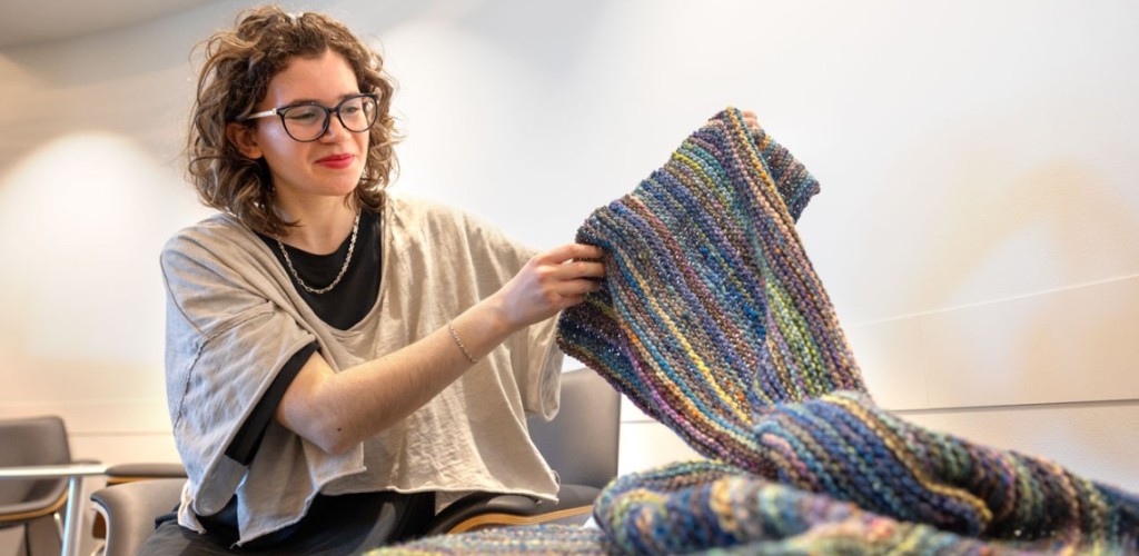 A woman holds up a long rainbow knitted blanket.