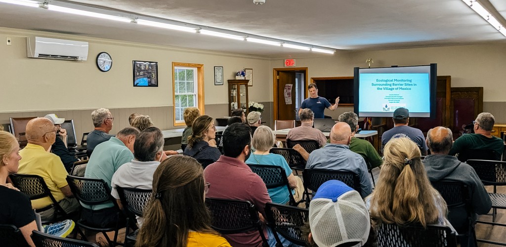 Photograph of a man standing at the front of a room with slides projected talking to about twenty people seated in rows.