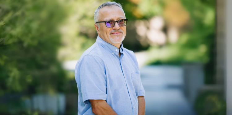 Toni stands in a shaded courtyard wearing a blue button-down shirt and glasses.