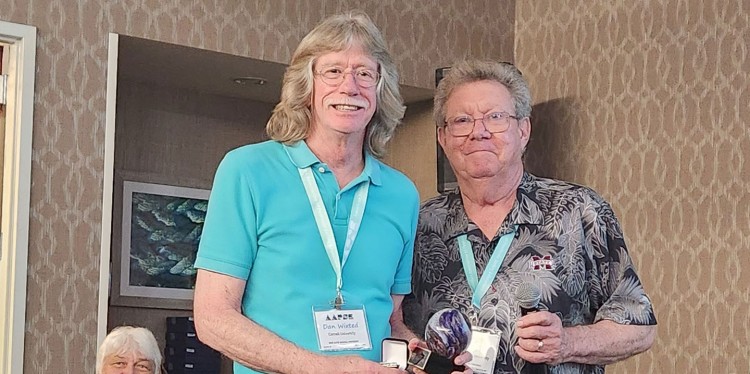Two people standing next to each other wearing name badges and an award, smiling for he camera. There is another person sitting behind them in the background 