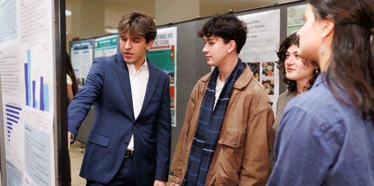 A student reads off a poster with three other students gathered around listening. 
