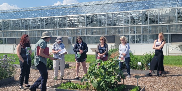 A person stands before a group, infront of a raised garden bed, with green houses in the background. 