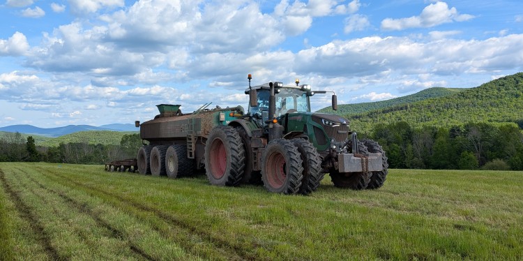 A large green tractor with a manure spreader stands in a green field, surrounded by rolling hills and a partly cloudy sky.