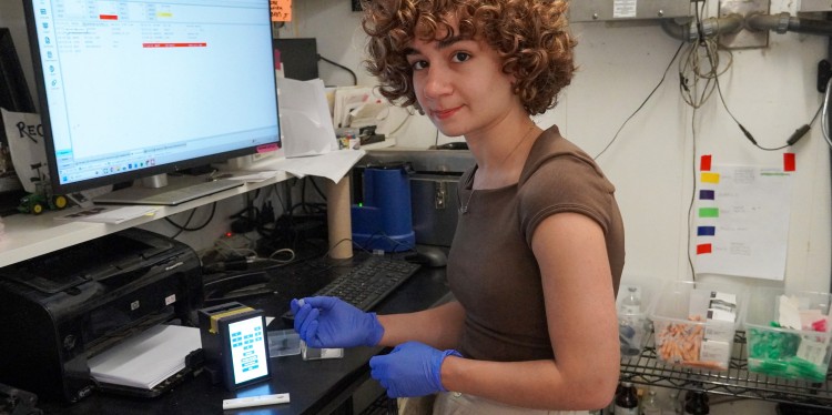 a woman wearing rubber gloves holds a plastic cassette is about to put it into a small square piece of tech with a touch screen