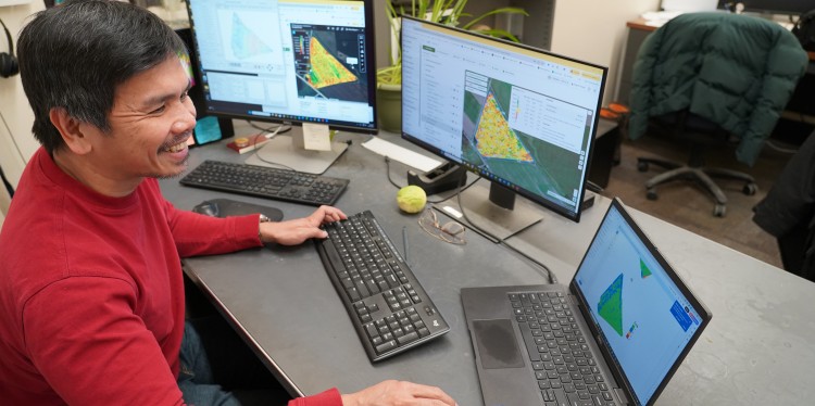 A man works on three computers. He is wearing a red shirt, sitting at a desk and smiling.