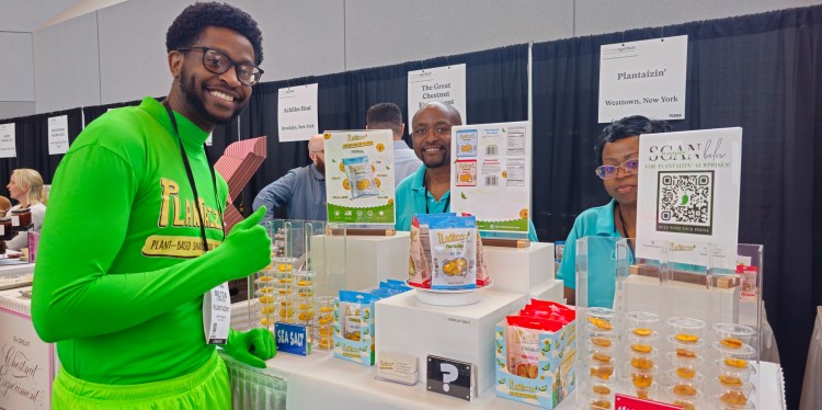 Three people, one of whom is wearing a green body suit, standing at a trade show booth. 