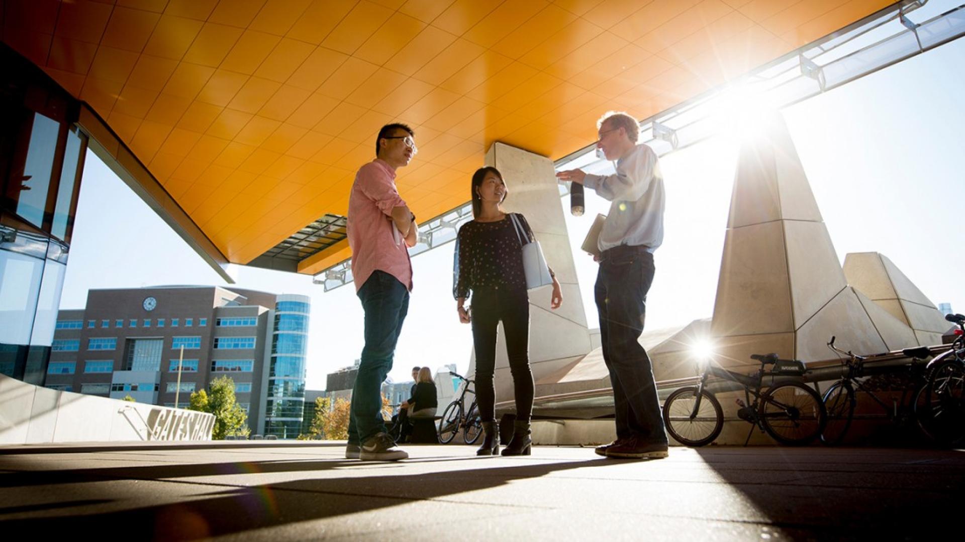 A woman and two men standing outside under a building overhang and talking