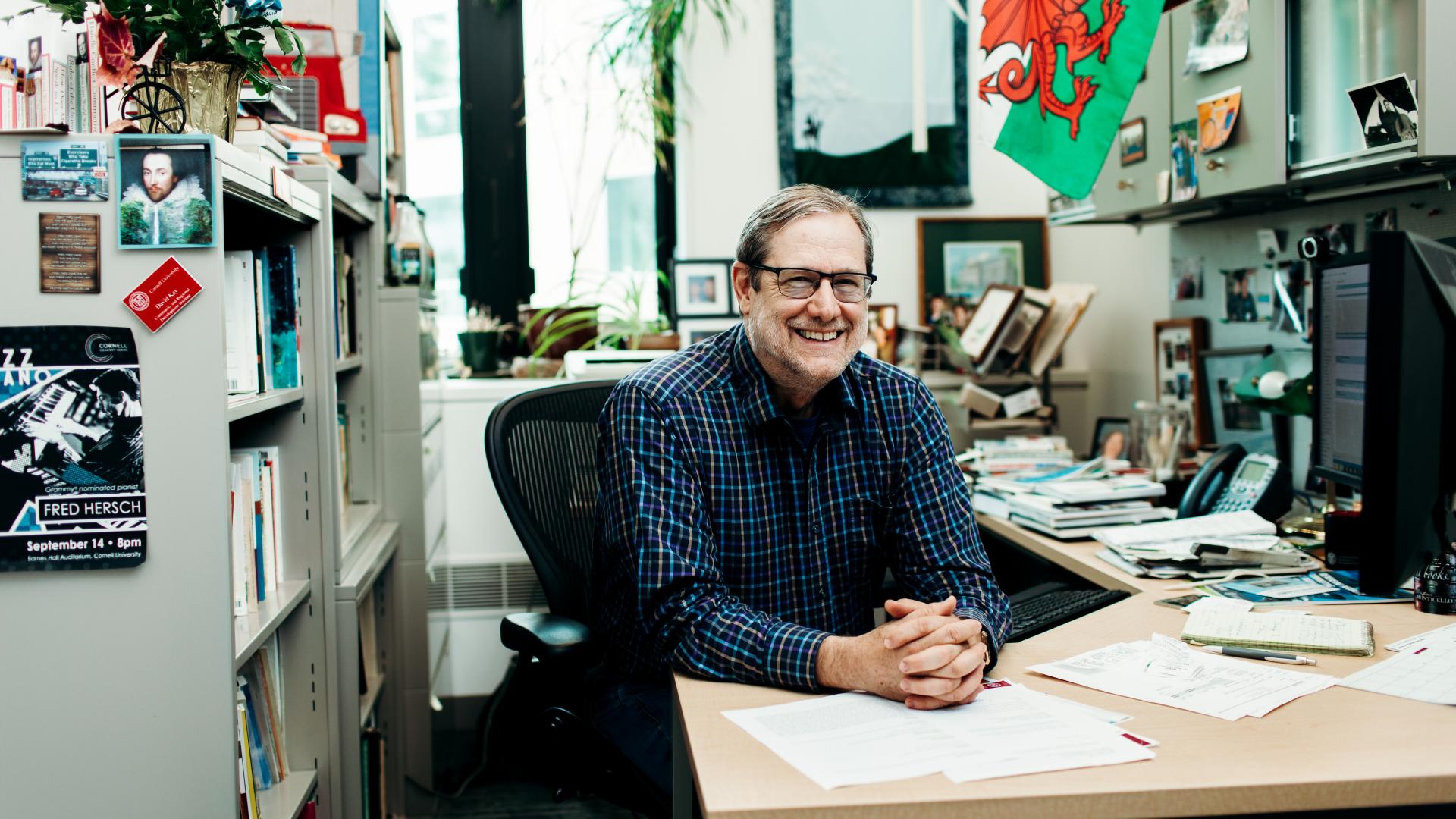 A man poses happily in an office while sitting with hands clamped on a desk