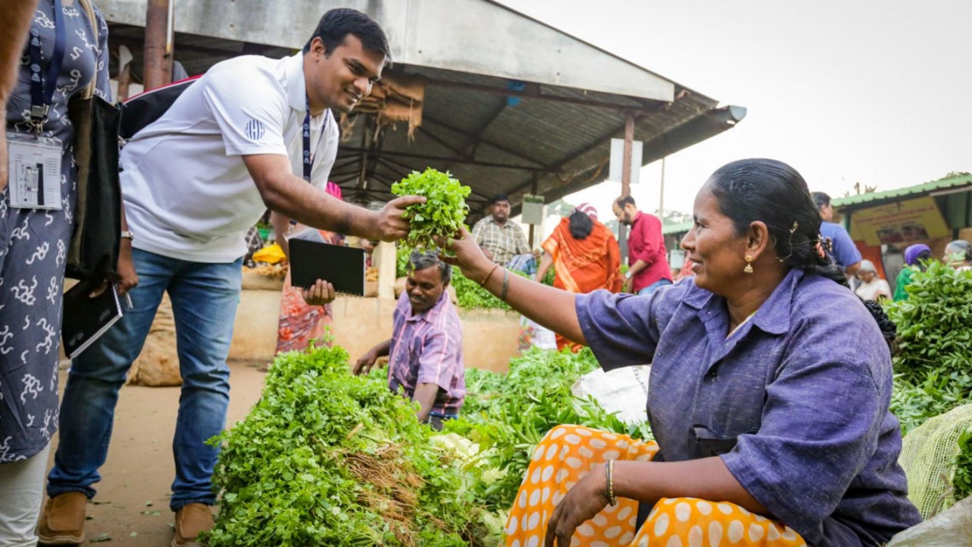 A man standing and taking leafy, green produce from a woman sitting in a market