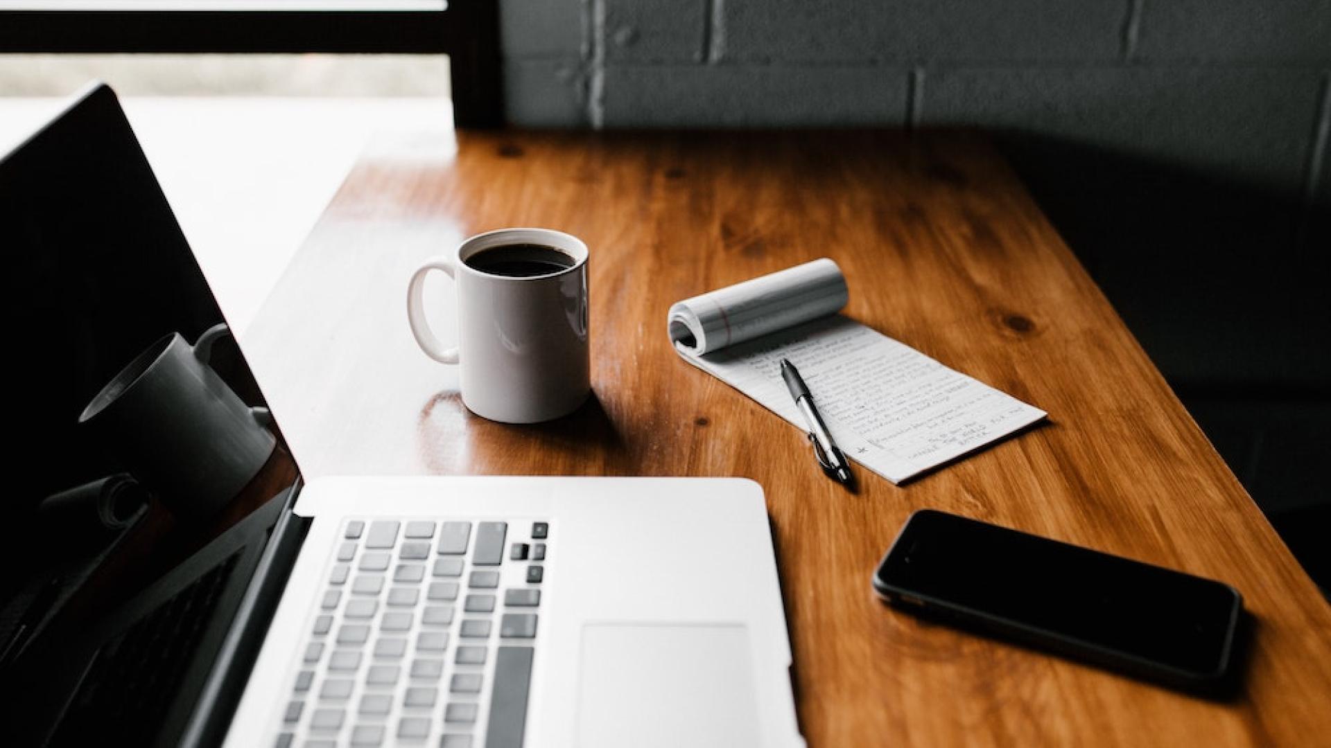 A laptop on a wooden table with a notepad and pen and a cup of coffee