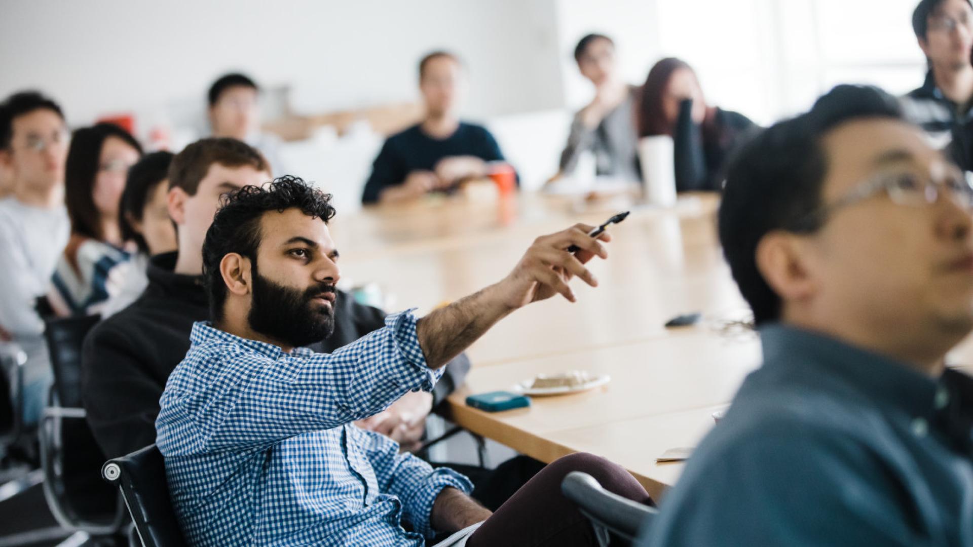 A man sitting at a table and gesturing at something out of frame. Other people are sitting at the table with him