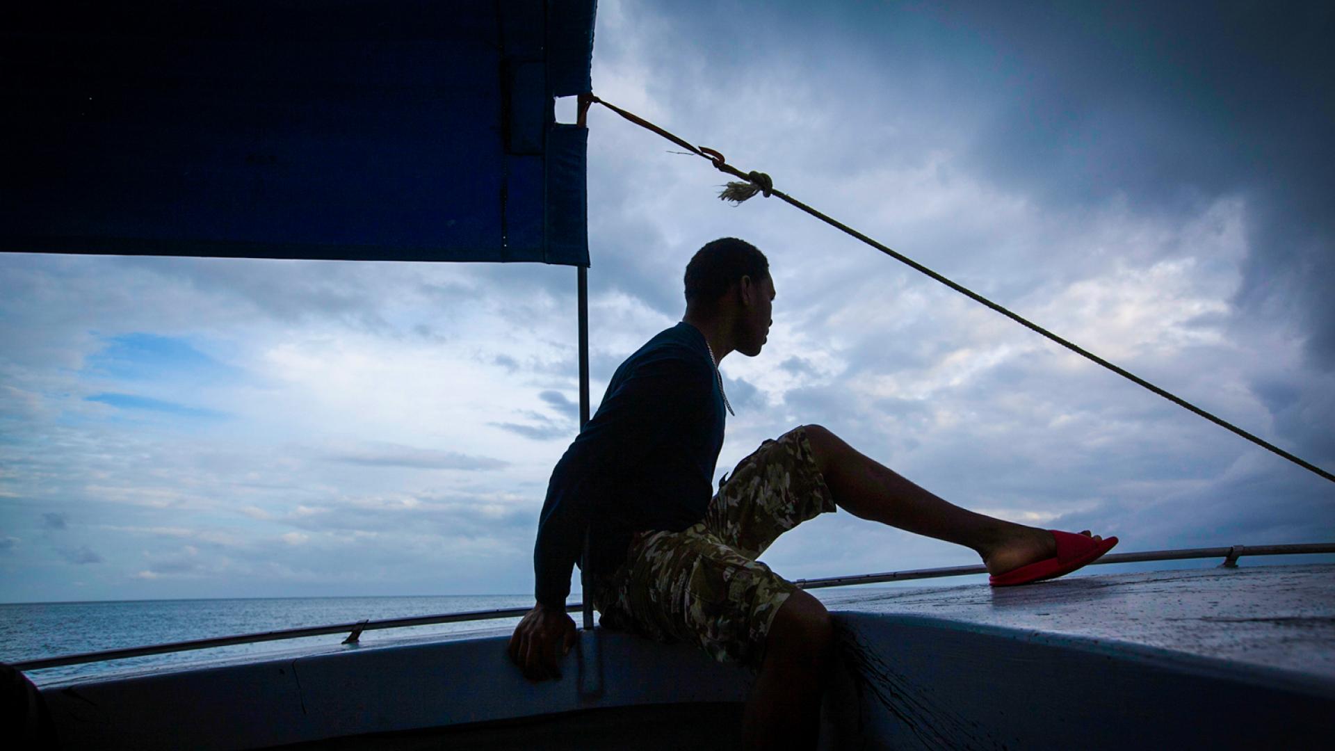 A person in silhouette at the front of a boat with a sail behind