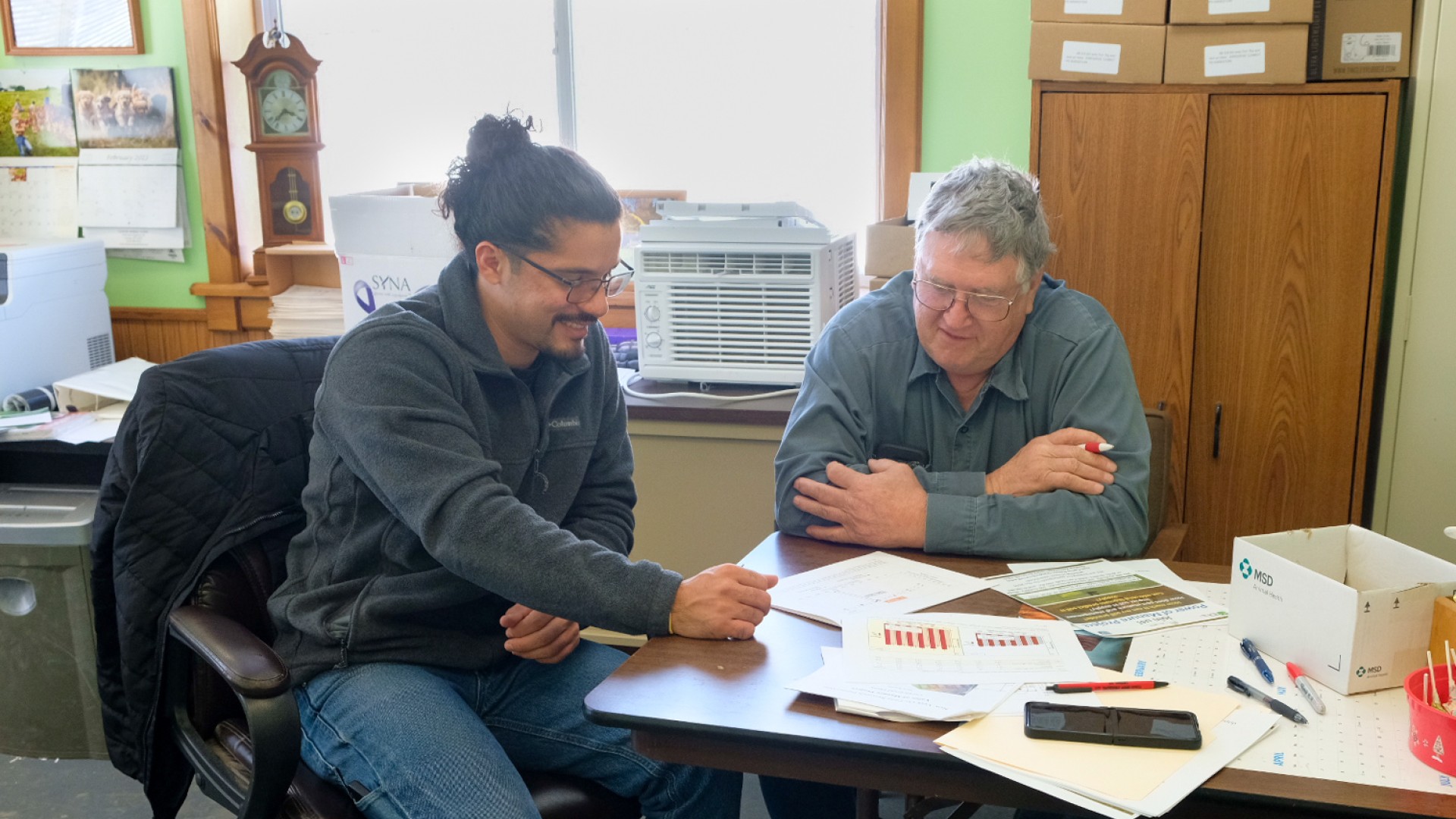 Two men sitting at a desk with papers on it talking.