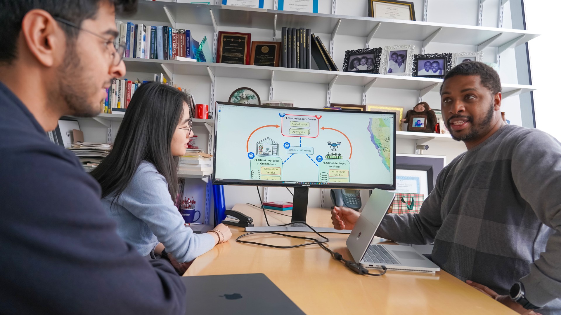 two men and a woman sit at a table talking with a schematic of farm and data on a view screen next to them