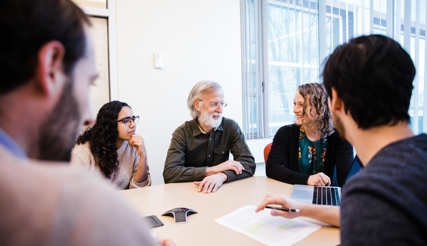 a man with white hair and a beard talks to a group of students at a table
