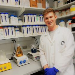 A man in a labcoat stands in front of lab shelves full of bottles