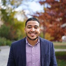 portrait of a man in a jacket and pink shirt in front of trees on the quad